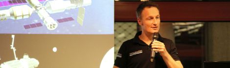 ESA-Astronaut Matthias Maurer hält einen Vortrag. Im Hintergrund sieht man auf seinen Vortragsfolien Raumstationen im Erdorbit und im Mondorbit.